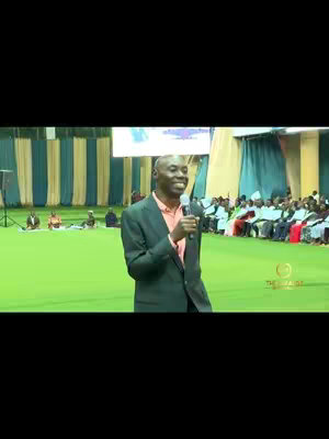 A man in a dark suit and pink shirt speaks into a microphone in front of a large crowd, with a green ground and a backdrop of white and blue curtains.