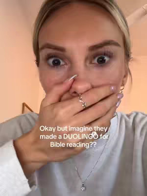 A close-up selfie of a woman with wide eyes and her hand covering her mouth in a surprised or shocked expression. The image features the woman's face prominently with a simple white background and a text overlay. She is wearing a white t-shirt and a delicate necklace.