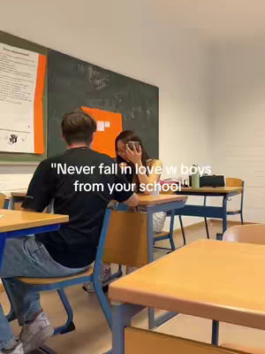 A classroom scene with two students, a male and a female, at desks. The male is towards the back of the frame, viewed from behind, while the female is facing the camera, obscured by her phone. The background includes a chalkboard and school desks.