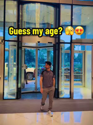 A man in a grey t-shirt and pants is walking towards the viewer. He is in front of a glass revolving door, with the interior of the building visible. The text overlay says "Guess my age?" and has emojis.