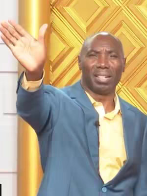 A medium shot of a Black man in a blue suit jacket, with a yellow shirt underneath, raising his right hand with his palm open. His expression is serious, with a slight furrow in his brow and slightly open mouth. The background is a golden, geometric pattern, giving a studio-like feel.