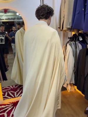 A man in a cream-colored garment stands with his back to the camera, looking into a mirror. The mirror reflects his image and two other people. The background shows clothing racks and neutral-toned suits.