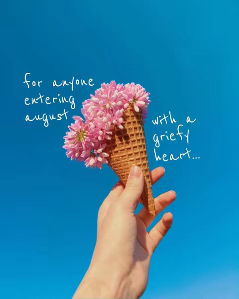 A hand holding a flower-filled ice cream cone against a bright blue sky. The cone is made of waffle, and the flowers are pink and arranged in a cluster.