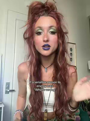 A young woman with striking makeup featuring green eyeshadow, purple lipstick, and red hair. Her hair is styled up with some hair hanging freely in curly waves. She wears a white tank top and various necklaces and bracelets and the background setting appears to be an indoor setting with a white door visible behind her.