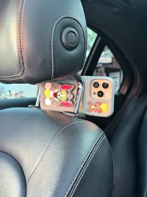 The image shows the back of a car seat with a phone mounted to the headrest. The phone is in a decorative case with cartoon characters. The mount also features cartoon characters.