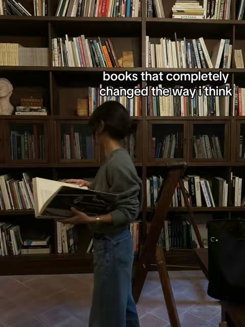 A woman standing in front of a large bookshelf. The background is filled with books on shelves. The woman is holding a large book that is open, the text "books that completely changed the way i think" is overlaid on the image.