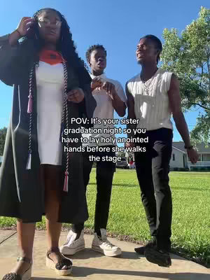 The image shows three young adults, two men and one woman, posing outdoors on a sunny day. The woman, in the center, wears a white dress and a graduation gown, with a colorful stole around her neck. The two men are on either side of her, one in a vest and the other in a white t-shirt under a jacket. The background suggests a residential area with green lawns and houses.