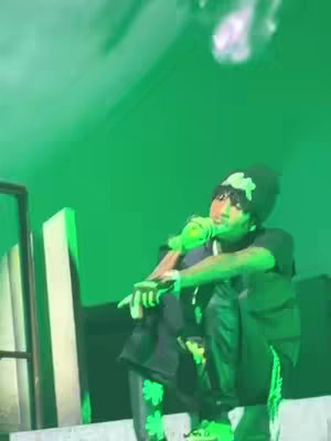 A person is on a stage, lit by green lights. The person is wearing a black beanie with a green shamrock on it, a black top, black pants with shamrocks down the side. The person is holding a green microphone. The background is green, and there is a hint of some foggy shapes at the top of the image.