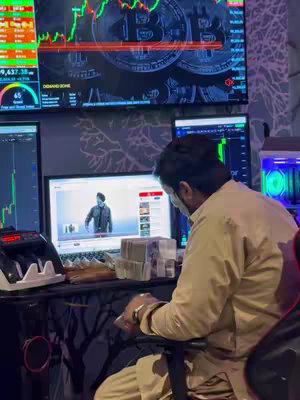 A man sits at a desk in a room with multiple computer screens. One screen shows a video of the man, another has a stock chart, and a third shows a currency amount. Stacks of cash are visible on the desk. The background has a decorative wall pattern and dim lighting.