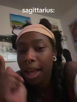 A young woman with dark skin, wearing a light pink headband, various piercings in her nose and lip, and braids. Text overlay at the top says 'sagittarius:'. She is looking at the camera and talking.