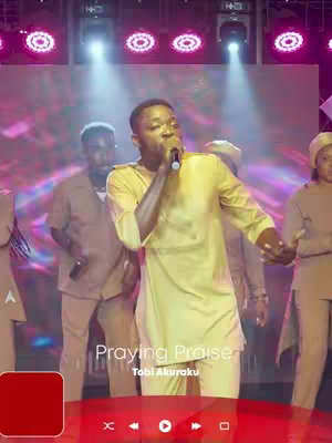 A cover image of a live gospel music performance. A Black man is in the center, singing into a microphone and wearing a long-sleeved yellow shirt. The background features a large stage and supporting singers. The text overlay at the bottom of the image in white font reads "Praying Praise" and "Tobi Akuraku".
