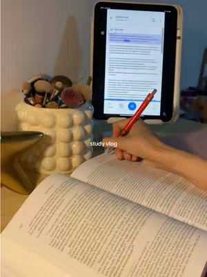 A person is studying. An open book and white tablet are presented in the foreground. The hand of a person is holding a red pen near the opened book. On the tablet is a document, with text highlighted in purple. Behind the tablet, there's a decorative jar filled with cosmetics.