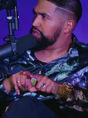 The image shows a close-up of a man wearing a dark floral-patterned shirt. He's looking slightly to his left, with a microphone situated near his mouth. His hands are visible, with the index fingers and thumbs of both hands touching. He's wearing a gold watch and a ring with a blue stone. The background is a solid purple color.