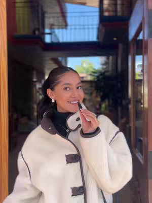 A woman stands in front of a doorway, looking at the camera and holding a lip product up to her mouth. She wears a white and brown jacket and has her hair pulled back. The background is a blurred interior.