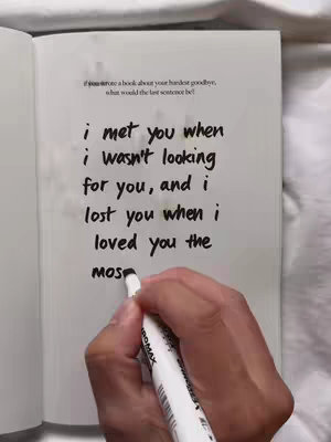 A hand holding a white pen writing on an open notebook. The notebook is white. The page contains a handwritten text in black ink. The text is a romantic quote.