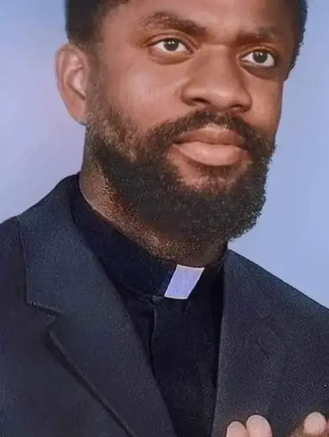 The image is a portrait of a man with a dark complexion, short, neatly-groomed dark hair, and a full, dark beard. He is wearing a black suit jacket over a black shirt with a white clerical collar. The background is a light, blurred blue. The man's expression is pleasant.