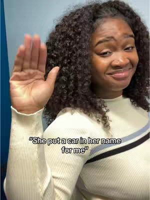A woman with curly dark hair raises her right hand as if to greet or gesture, her expression is slightly pained with a hint of a smile. She is wearing a white long-sleeved top with a line of dark blue and black trim near the neckline. Text overlayed on the image says, "She put a car in her name for me."