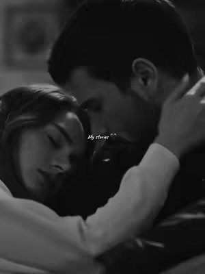 A black and white image of a man and a woman embracing. The woman is resting her head on the man's chest, and they appear to be hugging closely. The man has his arms around the woman. The photo suggests a romantic or intimate moment, with soft lighting and a blurred background.