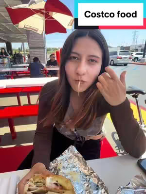 A woman sits at a table outdoors, holding a partially eaten sandwich in one hand, with a piece of food dangling from her mouth. The setting includes a red picnic table, a partially visible red and white umbrella, and other people in the background. The woman is looking directly at the camera, and there is text overlayed at the top of the image.