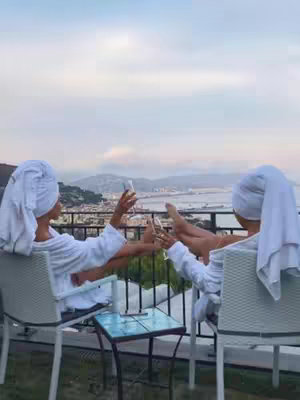 Two women in white robes and towels are sitting in white chairs on a balcony, clinking champagne glasses. They are overlooking a scenic view of a city and water in the background. The overall scene is one of relaxation and enjoyment.