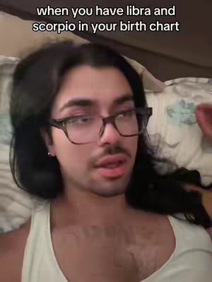 A person with dark hair and glasses is lying on a bed, looking towards the viewer. The background includes a patterned comforter. Text overlay reads 'when you have libra and scorpio in your birth chart'.