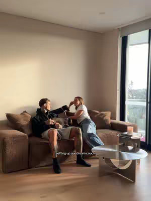 A photo shows a man and a woman sitting on a large, brown couch in a well-lit living room. The man is on the left, wearing a black hoodie and light-colored shorts, his legs covered in tattoos. The woman sits to his right, wearing a white t-shirt and blue jeans. They are facing each other, appearing to be in conversation. In front of them is a modern, low coffee table placed over a patterned rug. The background features a large window letting in natural light and a pale wall.