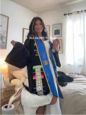 A young woman in a graduation gown with a blue and gold stole, holding onto someone out of frame. The image is set indoors with a bed and some framed photos in the background. The text overlay reads 'ALL GLORY TO GOD'.