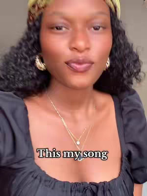 A close-up shot of a woman with curly black hair, wearing a black top, and gold earrings. She has a yellow and green patterned headband. Text overlay says "This my song". She is looking up diagonally with a subtle smile.