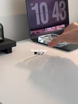 The image showcases a minimalist study setup on a white surface, likely a desk. A laptop is open with the time 10:43 AM displayed on the screen. A clear, rectangular object, perhaps a glass ashtray, is in front of the laptop near the center of the image. Other objects include what appears to be a dark-colored, small platform or stand and a small black container to the left. A hand appears to be moving the clear object. The background is plain and off-white.