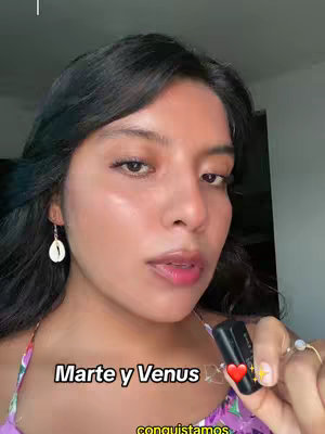 A selfie-style shot focused on a woman with dark hair, wearing a floral top and dangling shell earrings. The image is close-up, highlighting her face and upper chest. Overlay text near the bottom reads 'Marte y Venus'. The woman’s skin appears smooth with soft lighting. She holds a black object with her right hand.