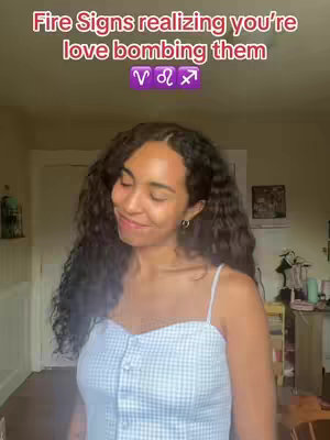 A young woman with long, wavy, dark hair is centered in the frame, smiling with closed eyes, wearing a light blue and white checkered dress with a buttoned bodice. The background shows indoor elements, suggesting a home environment. Overlayed text reads 'Fire Signs realizing you're love bombing them', featuring the astrological symbols for Aries, Leo, and Sagittarius.