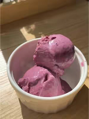 Two scoops of a vibrant pinkish-purple frozen dessert, possibly ice cream or sorbet, are presented in a small, white, slightly ridged bowl. The dessert has a smooth, glossy appearance with a slightly melting texture. The bowl sits on a wooden surface, with soft, warm lighting that highlights the texture of the dessert and the bowl.