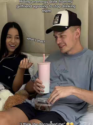 A couple is seated on a bed, with the man holding a pink and white ombre-colored cup and a small food container. Text overlays indicate the man is hiding the cup from his girlfriend. The woman is smiling and looking towards the man, while text overlays are on the top and bottom of the image.