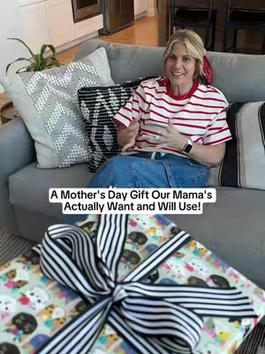 A woman is seated on a gray couch, smiling at the camera. She is wearing a red and white striped shirt and jeans. The background includes a gift wrapped in patterned paper, along with pillows and other home decor.