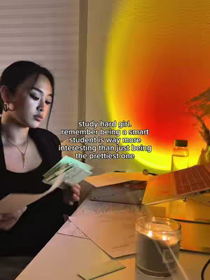 A woman is seated at a desk, engrossed in studying, with papers and a laptop spread out before her. The scene is softly lit with a setting sun lamp, creating a warm ambiance.
