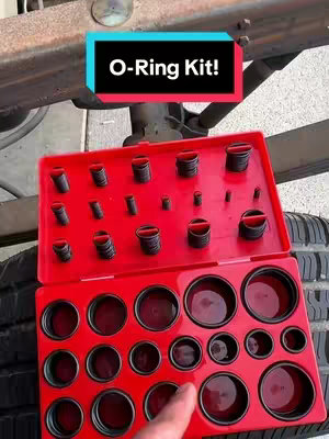 A close-up shot featuring a red tool kit with a variety of black o-rings, placed against the backdrop of a vehicle's undercarriage and tire. A text overlay in a blue rectangular box at the top reads 'O-Ring Kit!' 