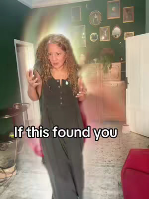 A medium shot shows a woman with curly graying hair standing in a room. She faces the camera and has both arms raised slightly with open palms. Behind her is a wall painted in dark green, adorned with various framed pictures. A bright, iridescent glow surrounds the woman. The text overlay "If this found you" appears at the bottom. The environment includes a glimpse of white doorway and a red couch. The overall composition blends spiritual elements with everyday surroundings.