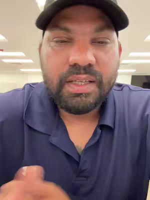 A man with a beard is centered in the frame, wearing a black cap and a dark blue collared shirt. The background is a brightly lit, nondescript space.