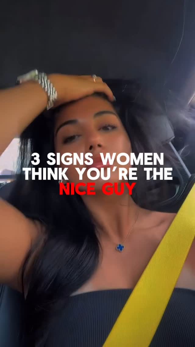 A woman with long dark hair is in the driver's seat of a car, looking at the camera. She is wearing a black top and a gold necklace with a blue charm. A yellow seat belt strap is visible. Text overlays read '3 SIGNS WOMEN THINK YOU'RE THE NICE GUY'. The background is blurred.