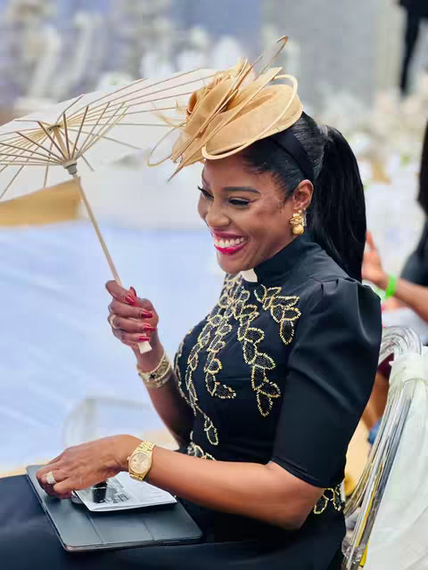 A smiling woman in a black dress with gold embroidery is holding a white and bamboo parasol. She wears a tan hat and earrings. Her expression is joyful while she is in an indoor, well-lit environment.