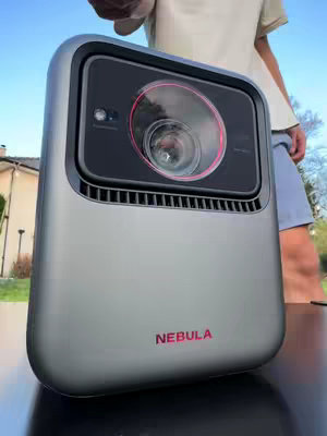 The cover image shows a metallic gray, rectangular projector with rounded edges. The lens has a black surround with a red inner circle. The word "NEBULA" is printed in red at the bottom. A person, with their arm and hand partly visible, appears to be holding the projector in front of a blue sky. The background features a house.
