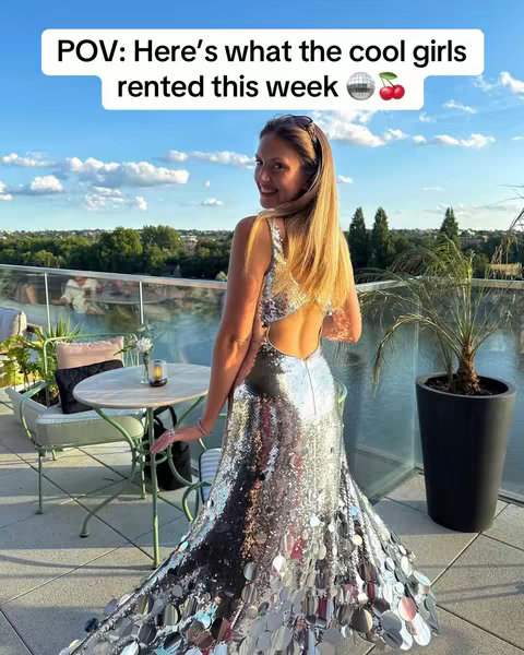 A woman in a sequined gown stands on a rooftop terrace, turning her head towards the camera with a smile. The background reveals a cityscape under a blue sky with fluffy clouds. A table with chairs and potted plants are in the foreground as well.