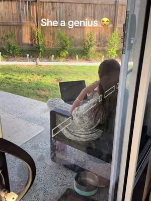 A person is sitting on a patio chair with a laptop and looking out to the yard through a glass door.