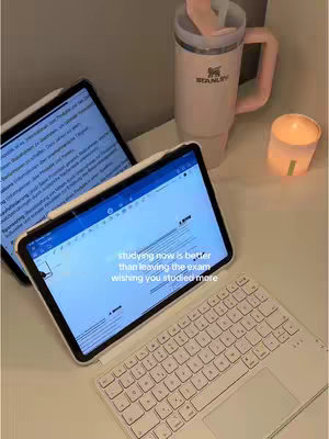 The image is a well-lit, top-down shot of a desk setup. Two tablets are open, displaying text, while a white keyboard rests in the foreground. A large tumbler and a small candle add to the aesthetic. The color palette is soft and warm, with whites and neutrals, creating a serene study environment. The composition is simple yet inviting.