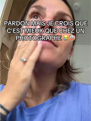 A woman with brown hair is visible from the chest up, her face a little bit to the right. She has a light skin tone and is wearing a blue shirt and a ring is on her left hand. Her left hand is raised to her face, covering her mouth and cheek. Text is overlaid at the top: "PARDON MAIS JE CROIS QUE C'EST MIEUX QUE CHEZ UN PHOTOGRAPHE".