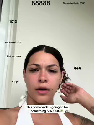 A selfie-style video cover image featuring a woman with dark hair against a plain background. The woman is looking directly at the camera with a slight expression of contemplation or concern. Text overlays with numbers and phrases, such as '88888', '1010', '444', 'You are FREEEEEE', 'Untouchable.', and ‘1111’ are arranged around her face. A text overlay at the bottom reads, 'This comeback is going to be something SERIOUS'.