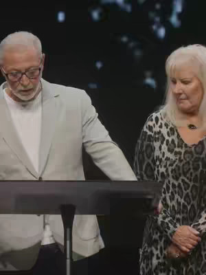 Two people, a man and a woman, are standing at a podium giving a speech. The man is on the left, wearing glasses and a light gray blazer over a white shirt. The woman on the right has blonde hair and is wearing a dress with a black and white animal print. They are both looking down and slightly to the left. The background is dark.