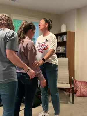 The cover image features three people in a room, with the focus on a woman in a white t-shirt and jeans. She's standing and embracing a younger person, whose back is to the camera. Another person is positioned to the left, partially visible. The overall lighting is natural and appears to be indoors. The background includes a window, a bookcase, and a chair.