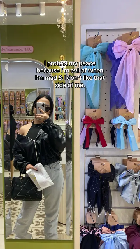 A woman in a black leather jacket and light wash jeans is taking a selfie in front of a mirror. The mirror reflects the interior of a shop, bow displays, and the text overlaying on her image.
