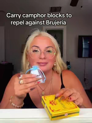 A woman with blonde hair and glasses holds a small blue bag and a yellow box with text, standing in front of a neutral background. The text overlay reads, "Carry camphor blocks to repel against Brujeria."